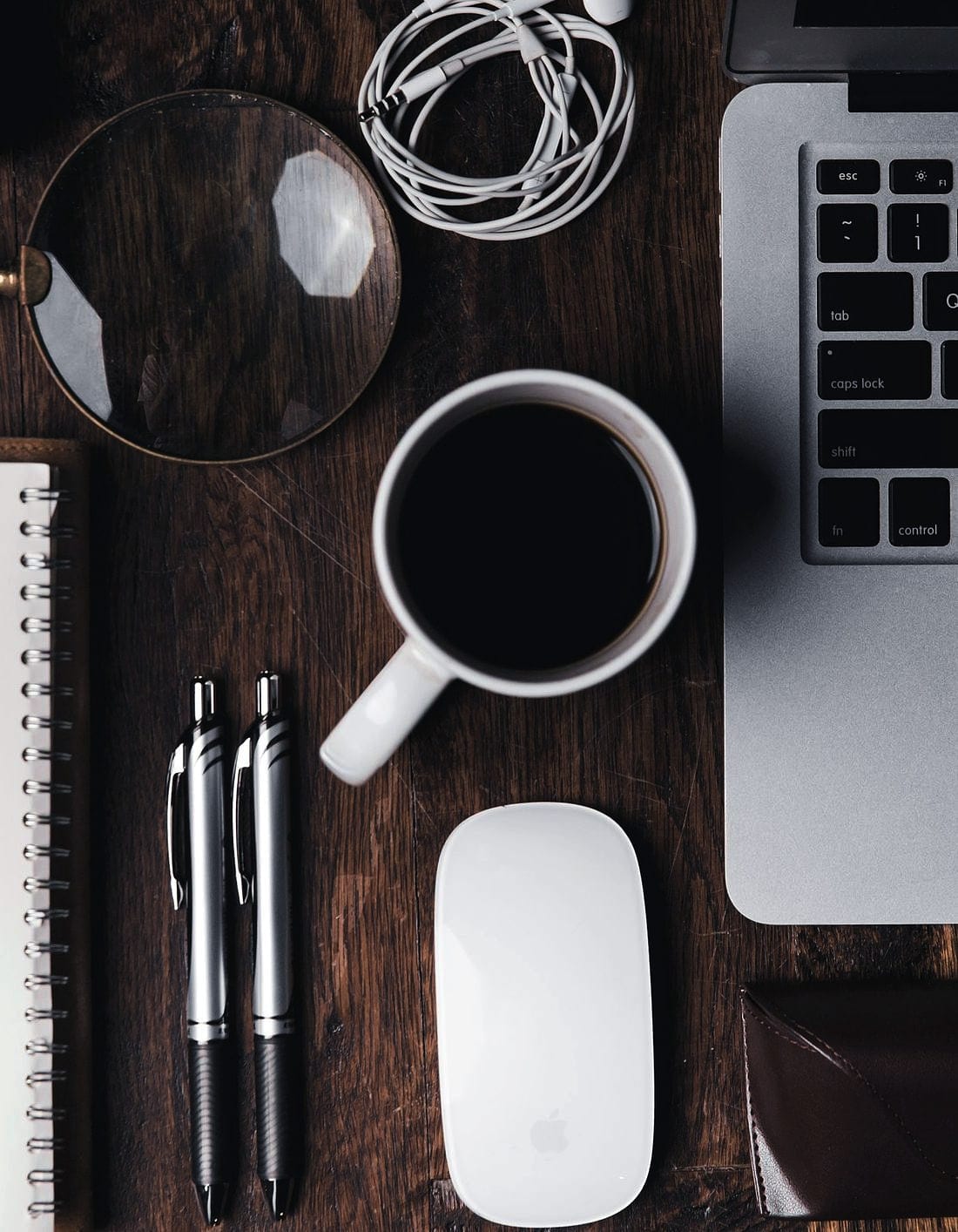 A magnifying glass on a desk between a notebook, a coffee and a laptop