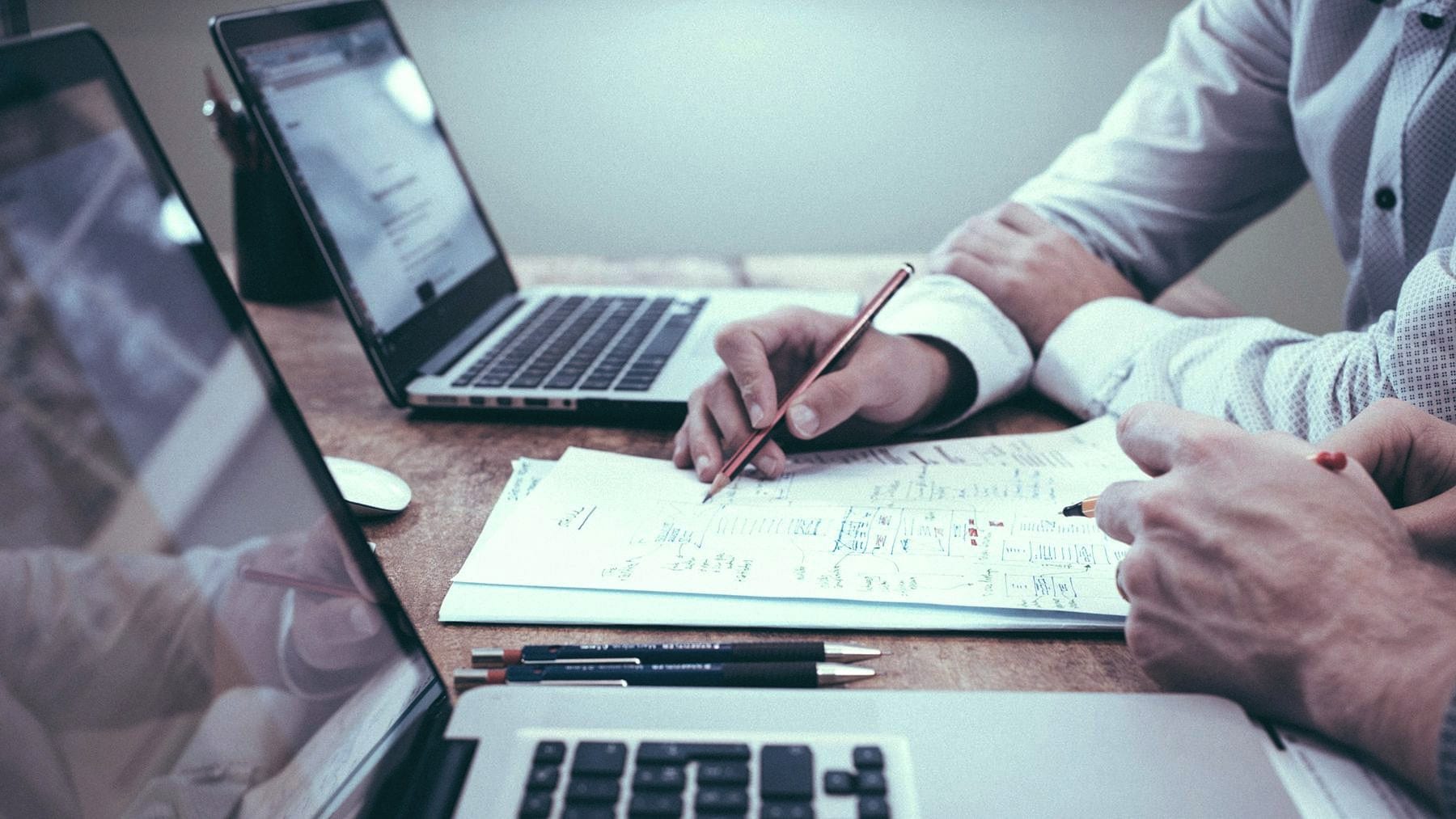Hands writing notes on paper beside open laptops